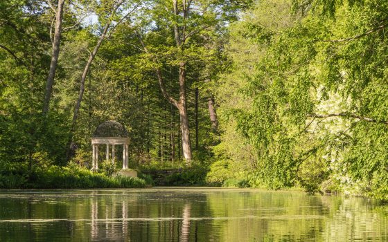 A small stone decorative structure is seen across a large lake surrounded by green trees