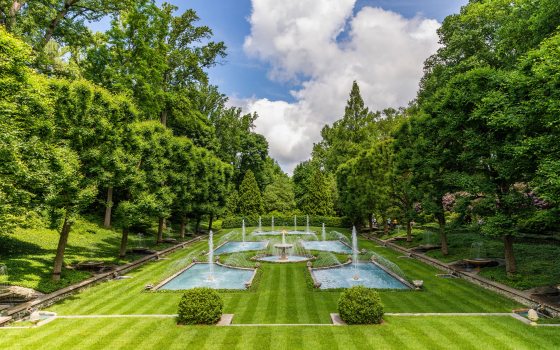 A long rectangular water garden of five pools and multiple fountains extends out in a large area surrounded by green trees under a blue sky