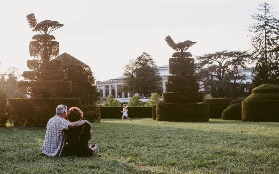 A couple sits on the ground surrounded by shaped topiary with a child running in the distance during sunset
