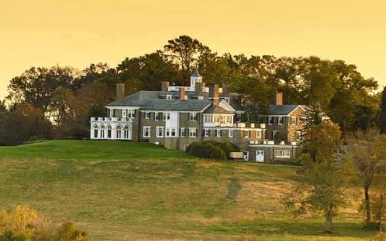 A mansion sitting at the crest of a hill, surrounded by trees and fields against a golden sky.