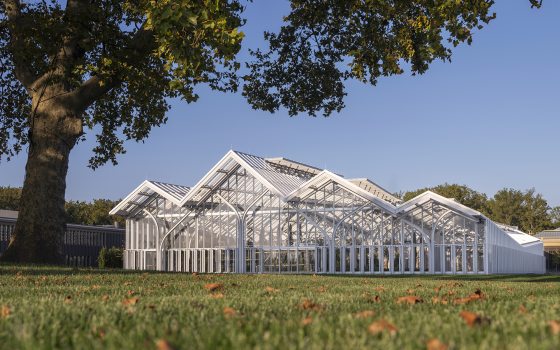 a glasshouse with a multi-peaked roof rises from a wide green lawn dotted with fallen autumn leaves