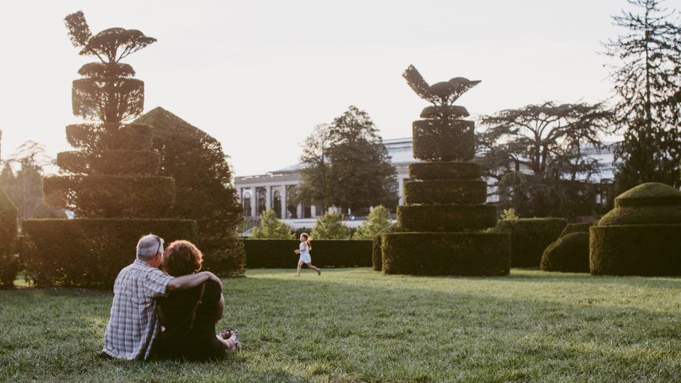 A couple sits on the ground surrounded by shaped topiary with a child running in the distance during sunset