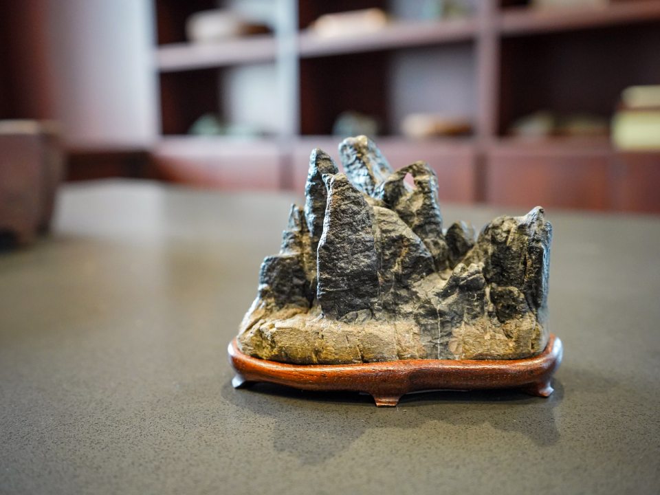 The alt text for this image should be:  A close-up, low-angle shot of a small, dark, craggy rock formation carving, resembling miniature mountains or peaks. It sits on a smooth, dark wooden stand with curved feet. The object is centered on a grey, textured table, with a blurred background of dark wooden shelving or cabinetry.
