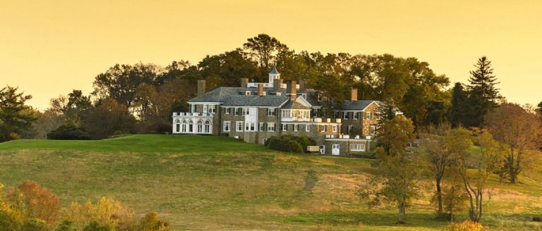 A mansion sitting at the crest of a hill, surrounded by trees and fields against a golden sky.