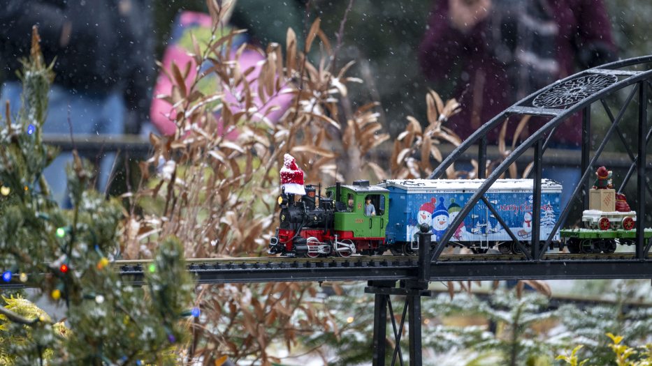 A model train featuring Santa Claus and holiday decorations runs on tracks through a snowy outdoor scene with onlookers in the background.