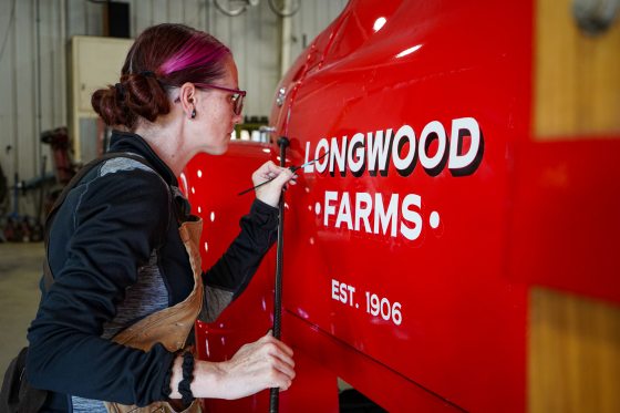 A person writing "Longwood Farms" with paint on a red pick-up truck.