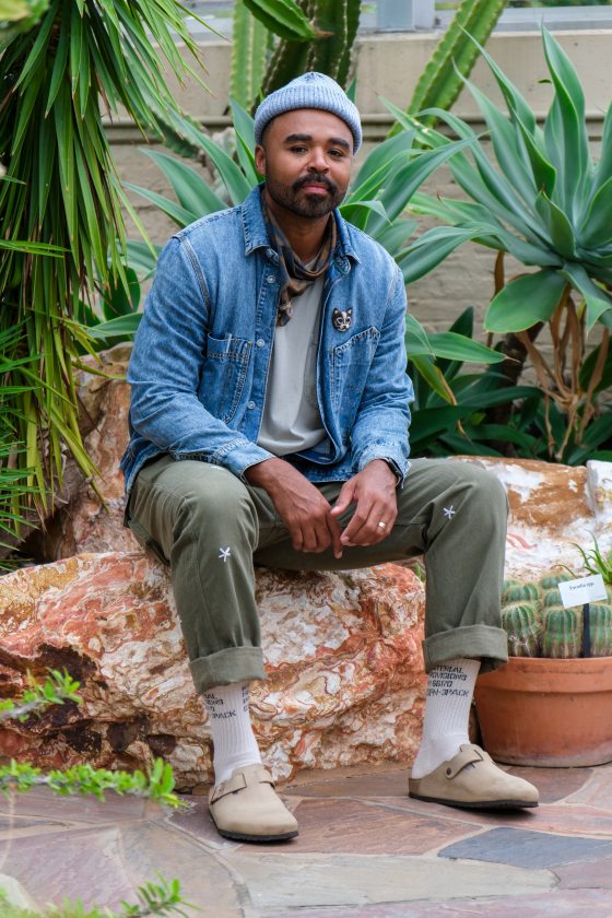 A person in a blue shirt, green pants, and hat with darker skin sitting on a large rock in a garden.