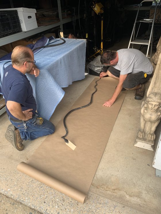 Two people in a metal shop on their knees examining a black iron snake.