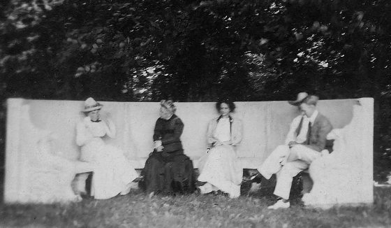 A historic black and white photo of four people sitting on a stone bench.