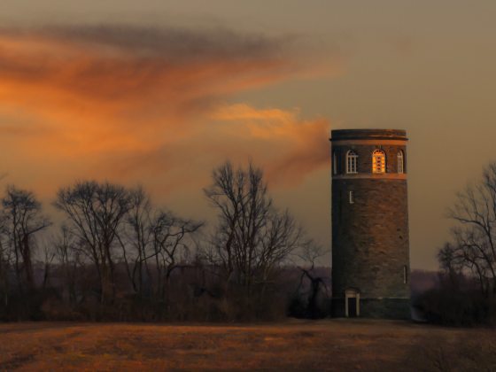 Clouds reflect an orange sunset over a field in winter, with leafless trees in the background and a stone tower on the right.