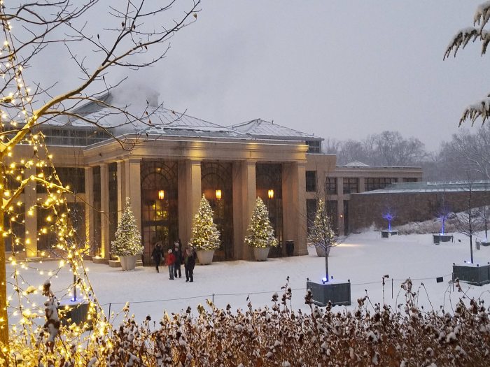 A glass and concrete conservatory is seen through snow flurries with a snow-covered ground and tress with Christmas lights