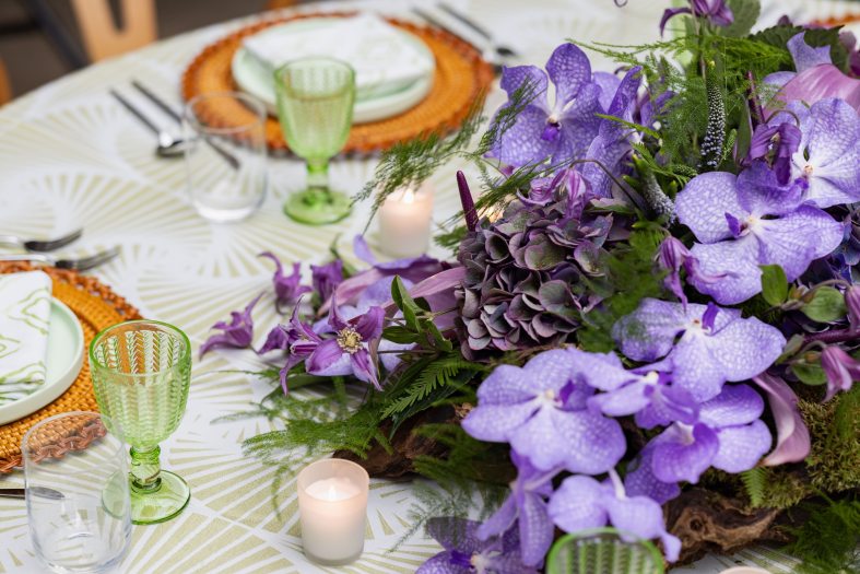 Closeup of table settings on a white and gold patterned tablecloth, with woven basket-like servers, round white plates, cloth napkins, green glass goblets, votive candles, and a floral arrangement in green and purple hues.